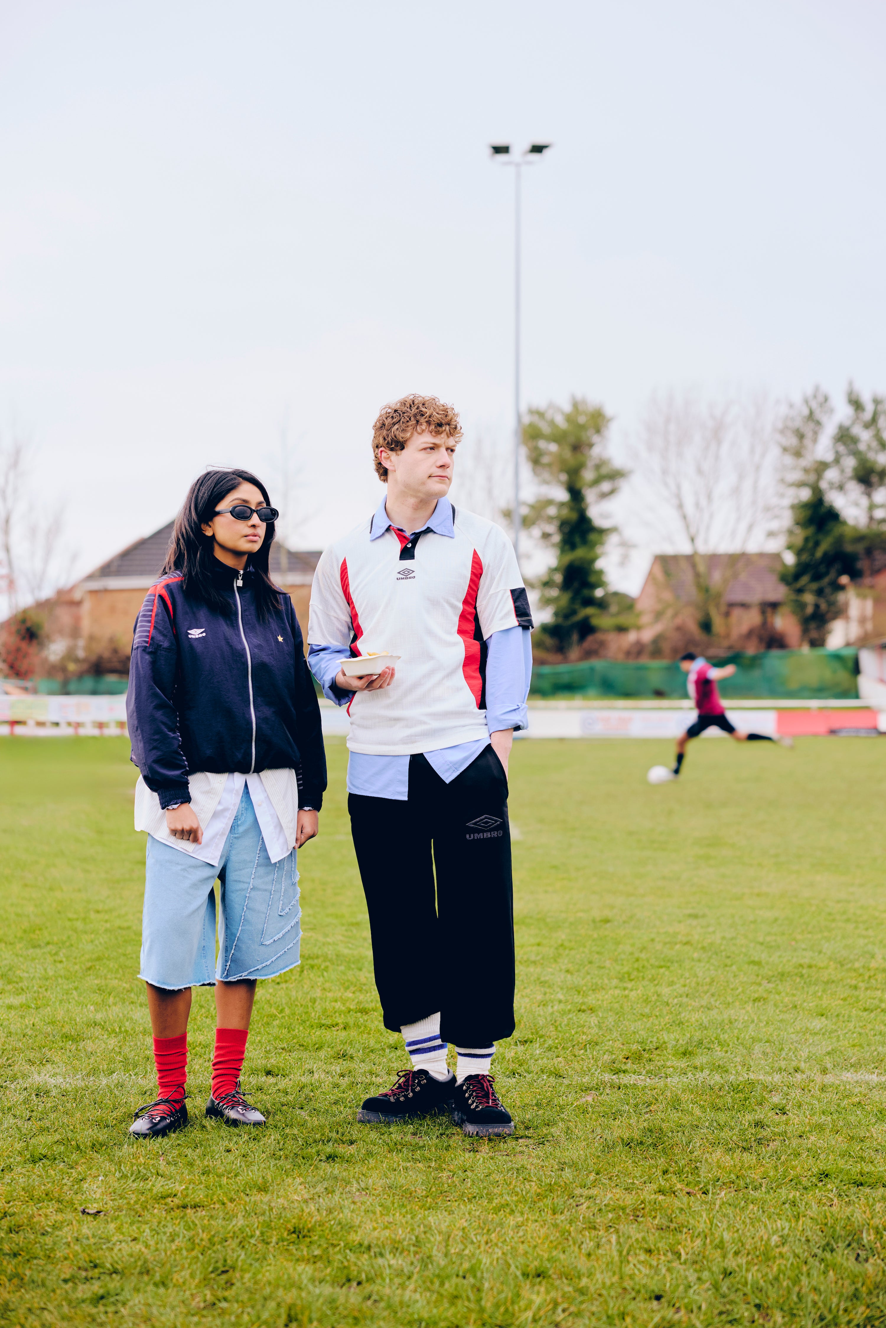 Two people, wearing retro Umbro, standing on a grassy field with a person in the background playing soccer.