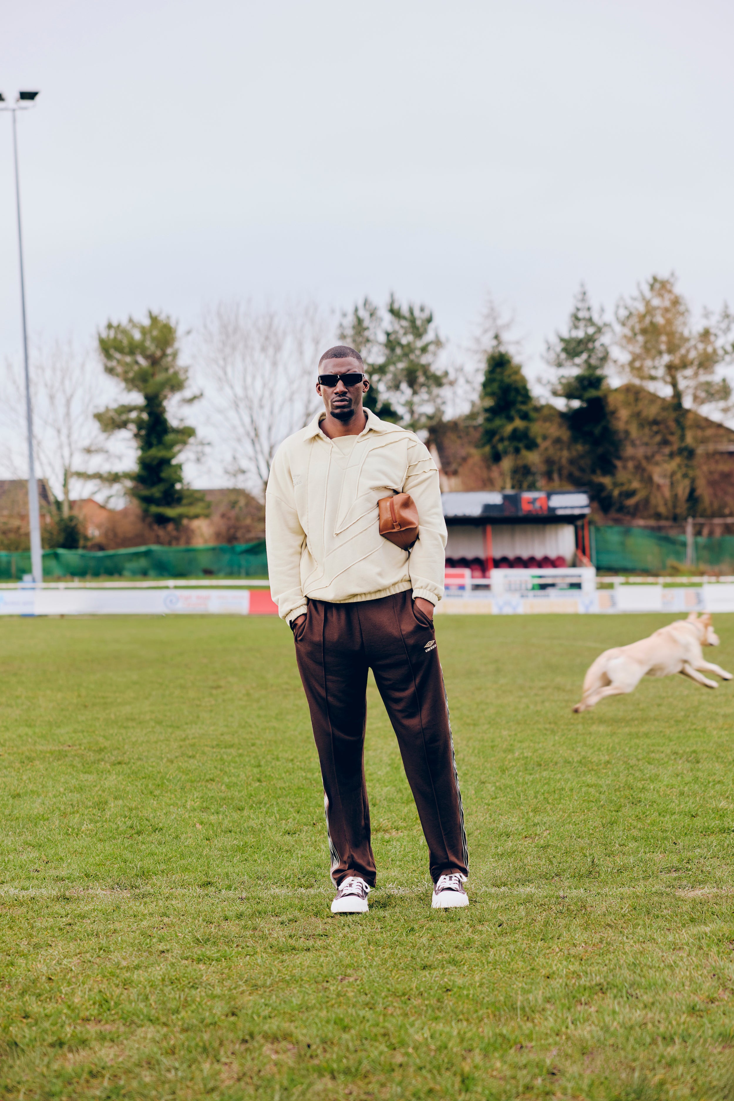 Man in sunglasses, wearing an Umbro top, standing on a grassy field with a dog in the background